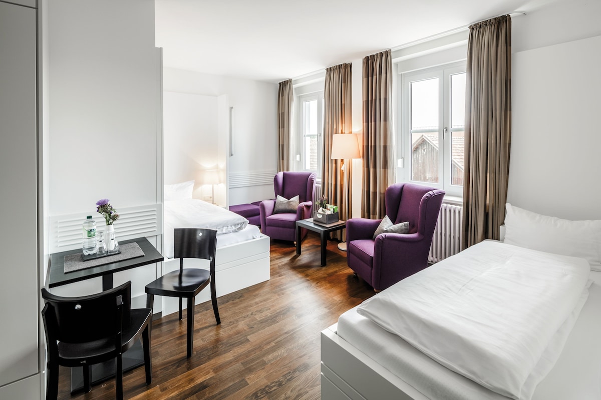 Een hotelkamer met een bed, twee paarse fauteuils, een tafeltje met een lamp, een bureau met stoel en een raam met gordijnen.