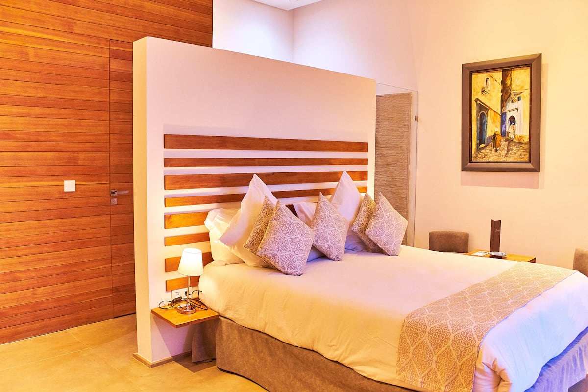A modern hotel room with a wooden panel wall, a large bed with patterned pillows, a bedside table with a lamp, and a framed artwork on the wall.