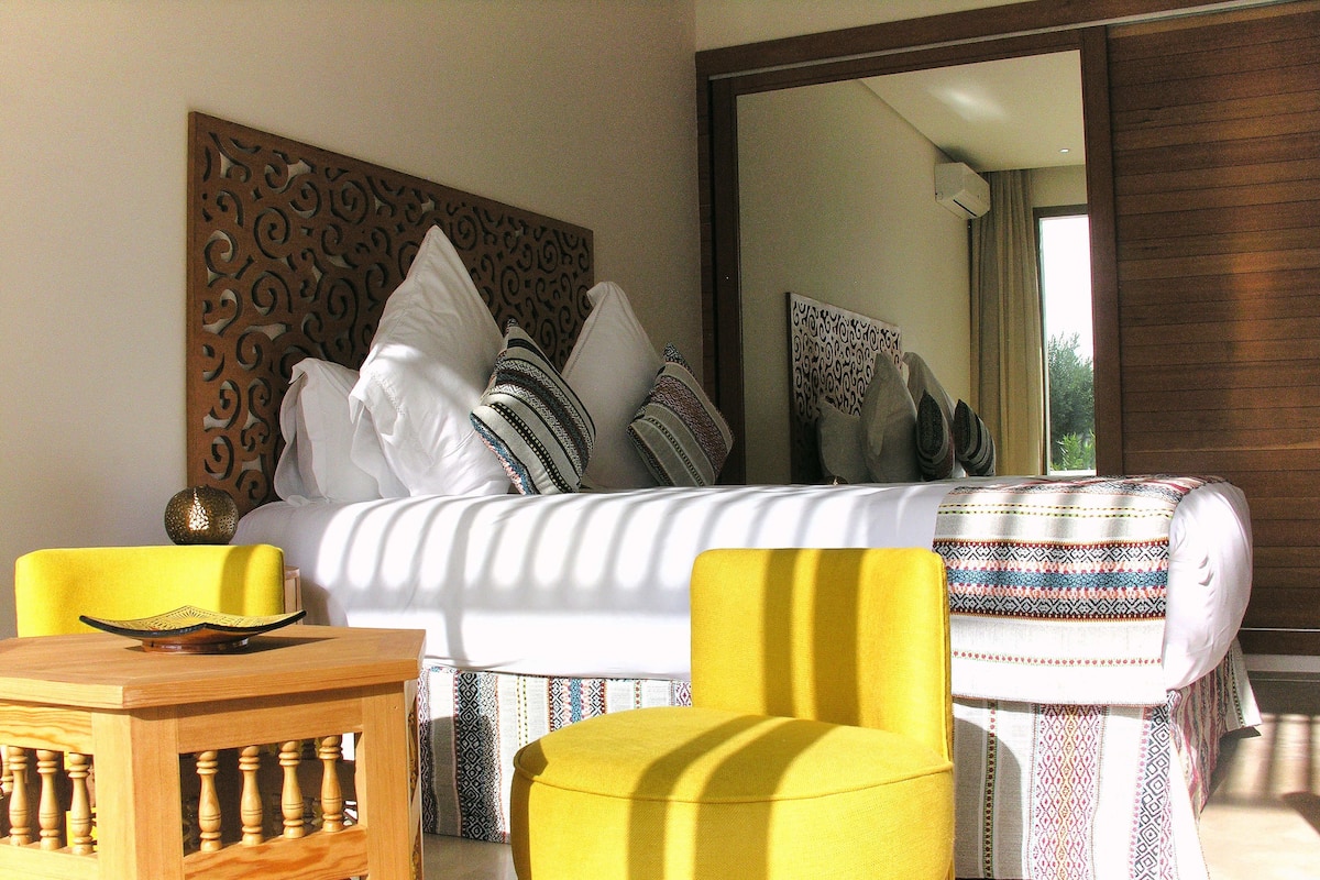 A bedroom with a bed, pillows, a yellow chair, a wooden side table, and a large mirror.