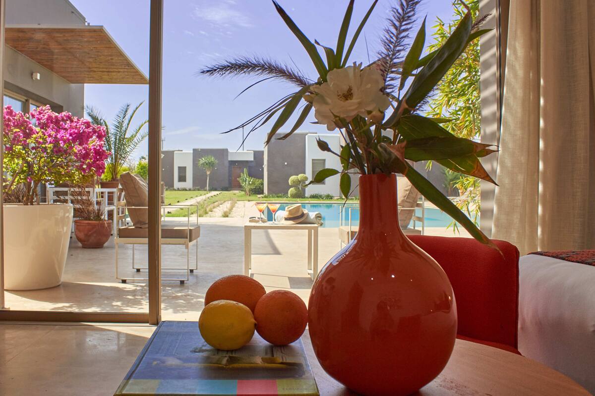 A room with a large window, a vase with flowers and oranges, a pool, and outdoor furniture.
