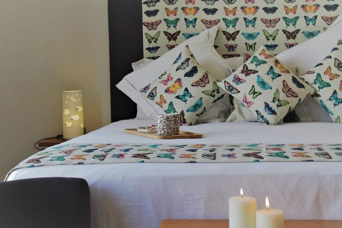 A neatly made bed with colorful butterfly-patterned pillows, a bedside table with a candle, and a decorative wall piece.