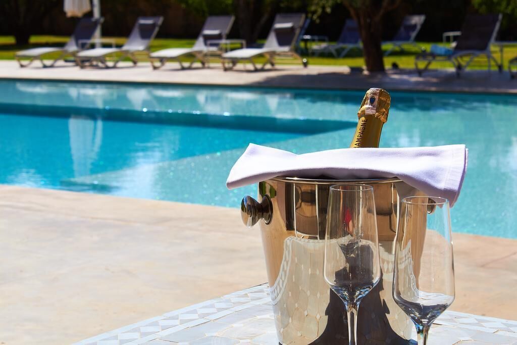 A poolside setting with a champagne bottle in a bucket, surrounded by empty wine glasses, and lounge chairs in the background.