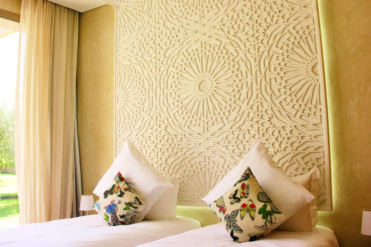 A hotel room with a decorative wall, two beds with floral pillows, and a window with curtains.