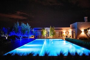 Vista nocturna de un área de piscina moderna con una pileta rectangular, rodeada de árboles y un edificio con iluminación cálida.