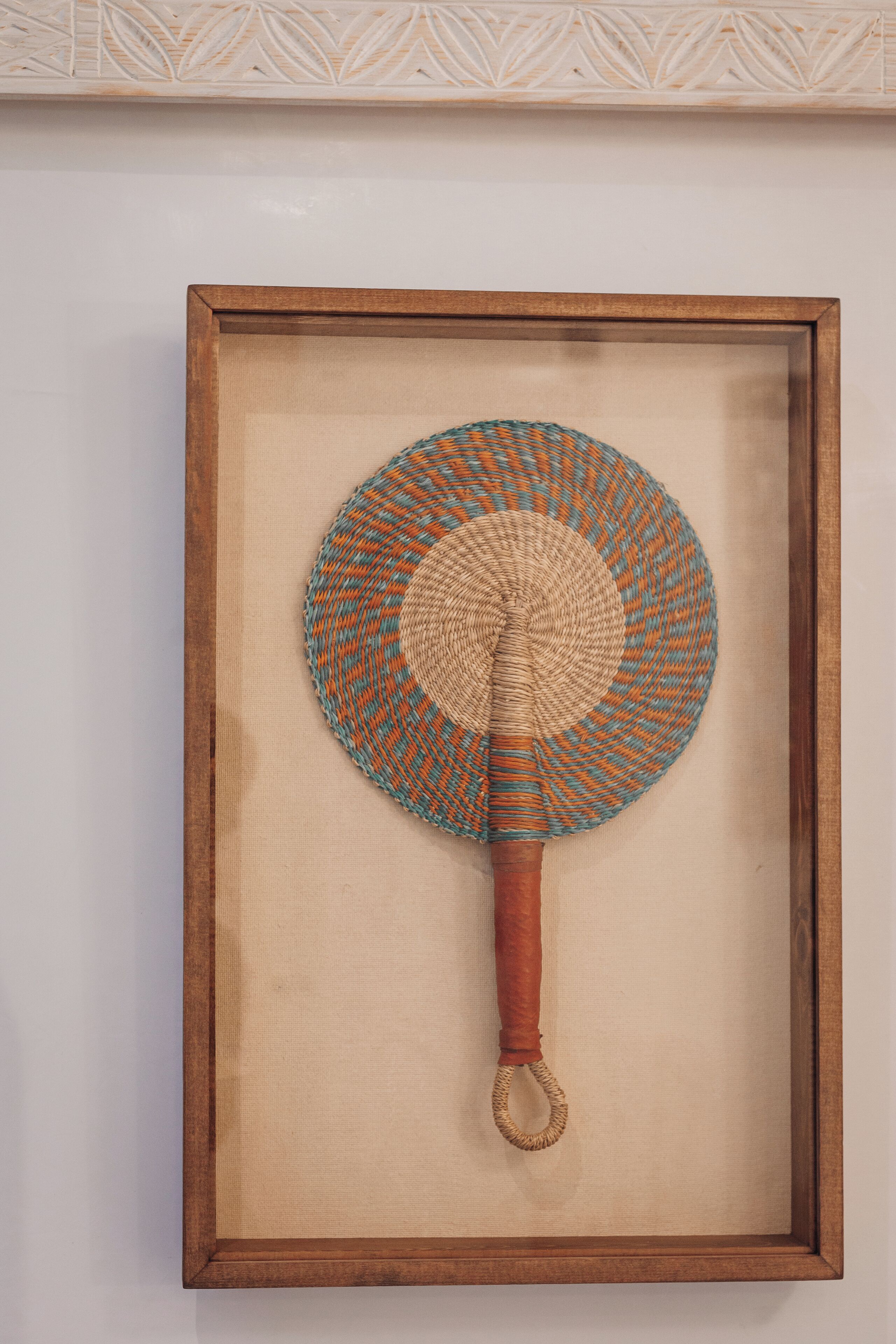 A framed woven fan with a wooden handle and a circular base, displayed on a wall.