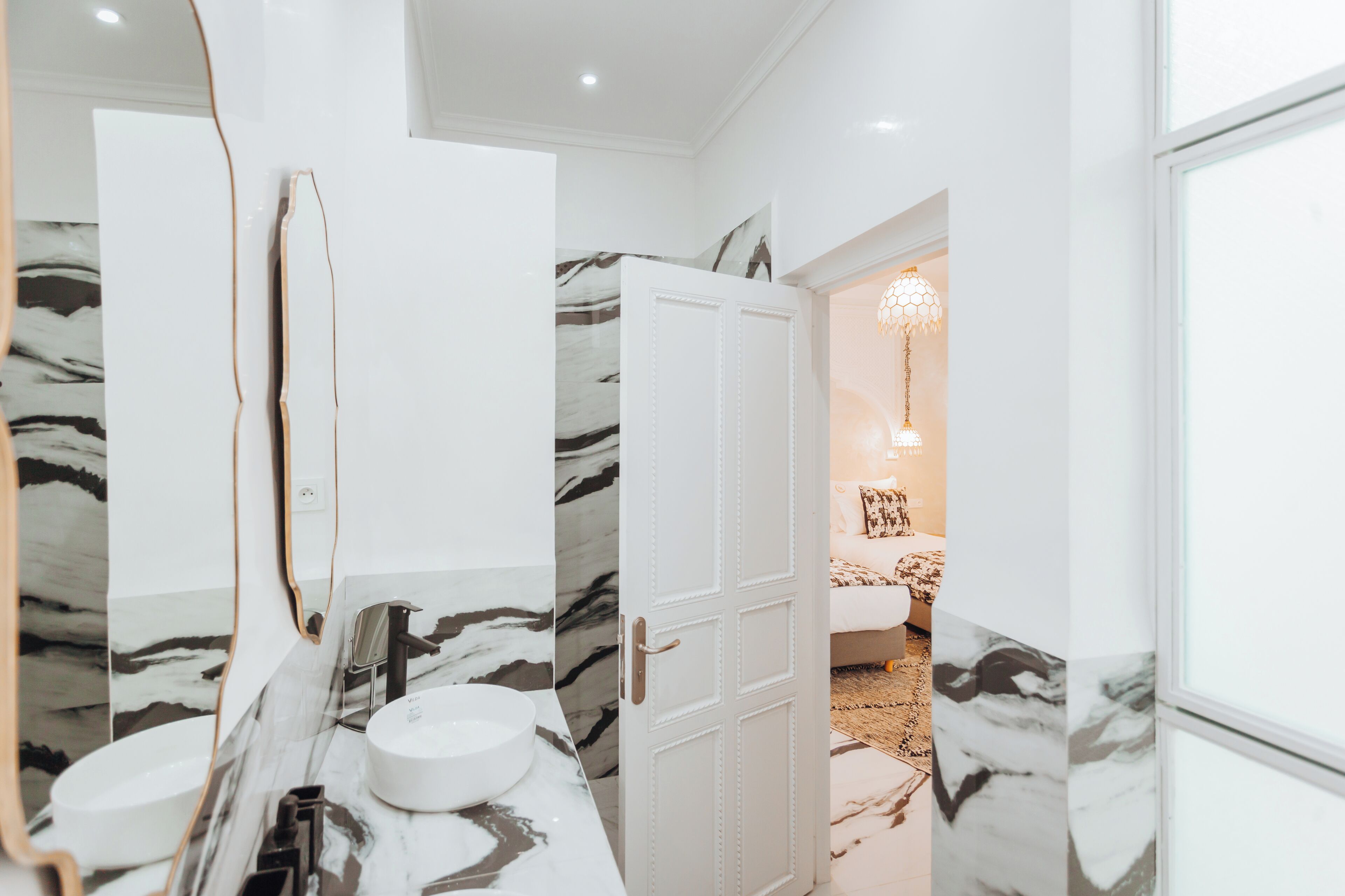 A bathroom with a marble vanity, a white door, and a view into a bedroom with a bed and a chandelier.
