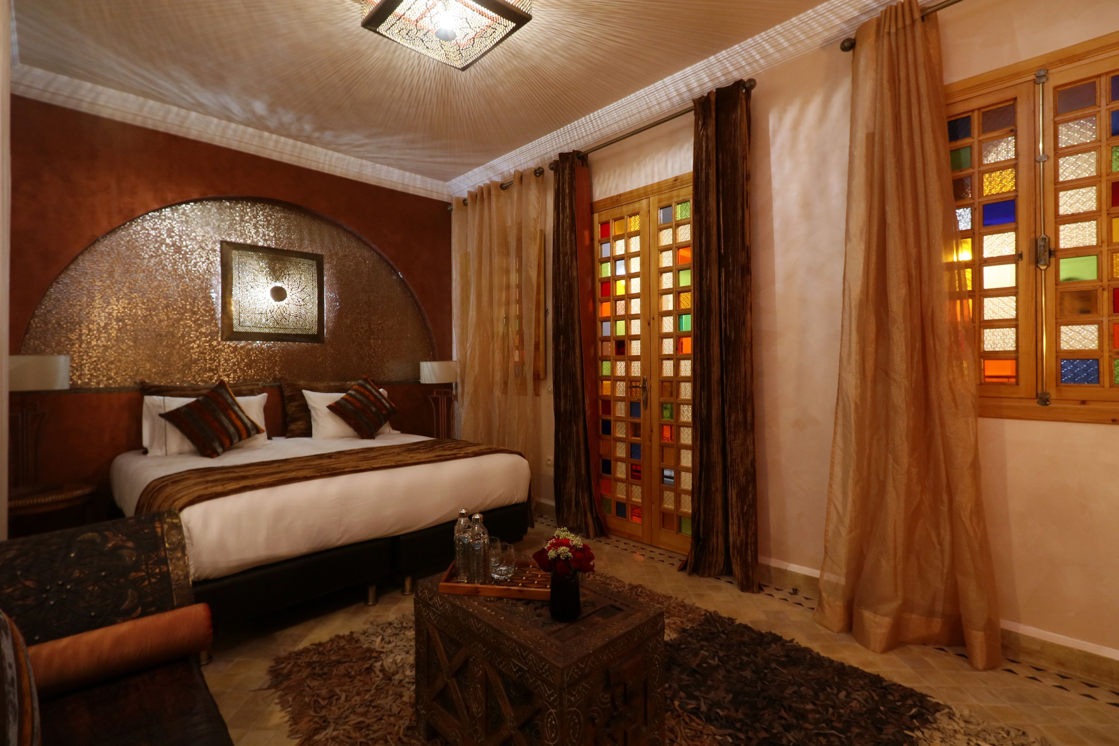 A spacious bedroom with a large bed, a wooden headboard, a decorative ceiling light, a window with stained glass, and a small wooden table with a vase of flowers.