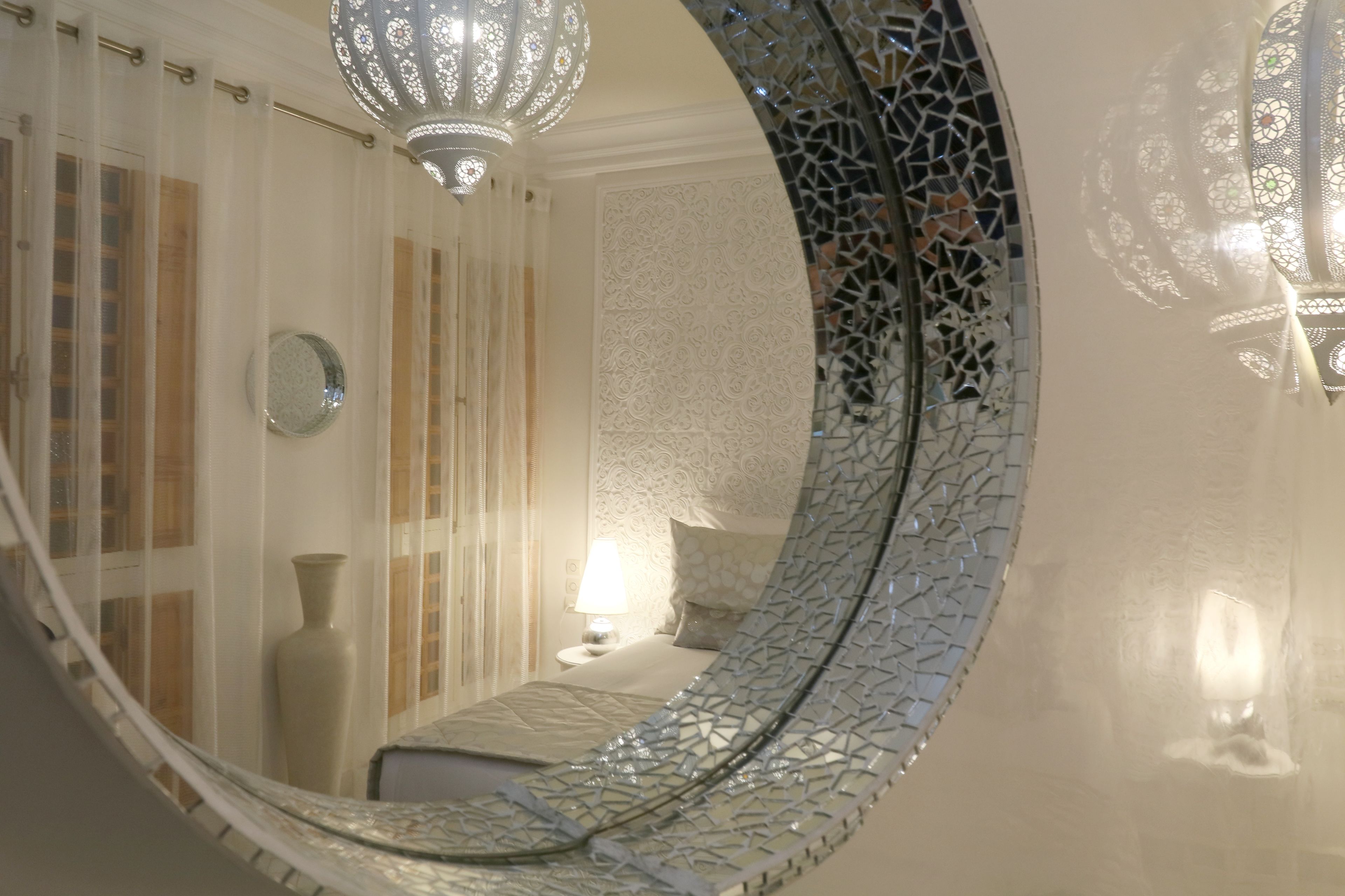 A circular mirror with intricate mosaic patterns, a bed with a white headboard, a bedside lamp, and a decorative vase.