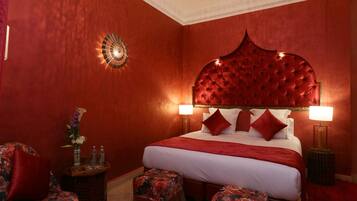 A red-themed bedroom with a tufted headboard, a large bed, two bedside lamps, a floral chair, and a small side table with a vase.