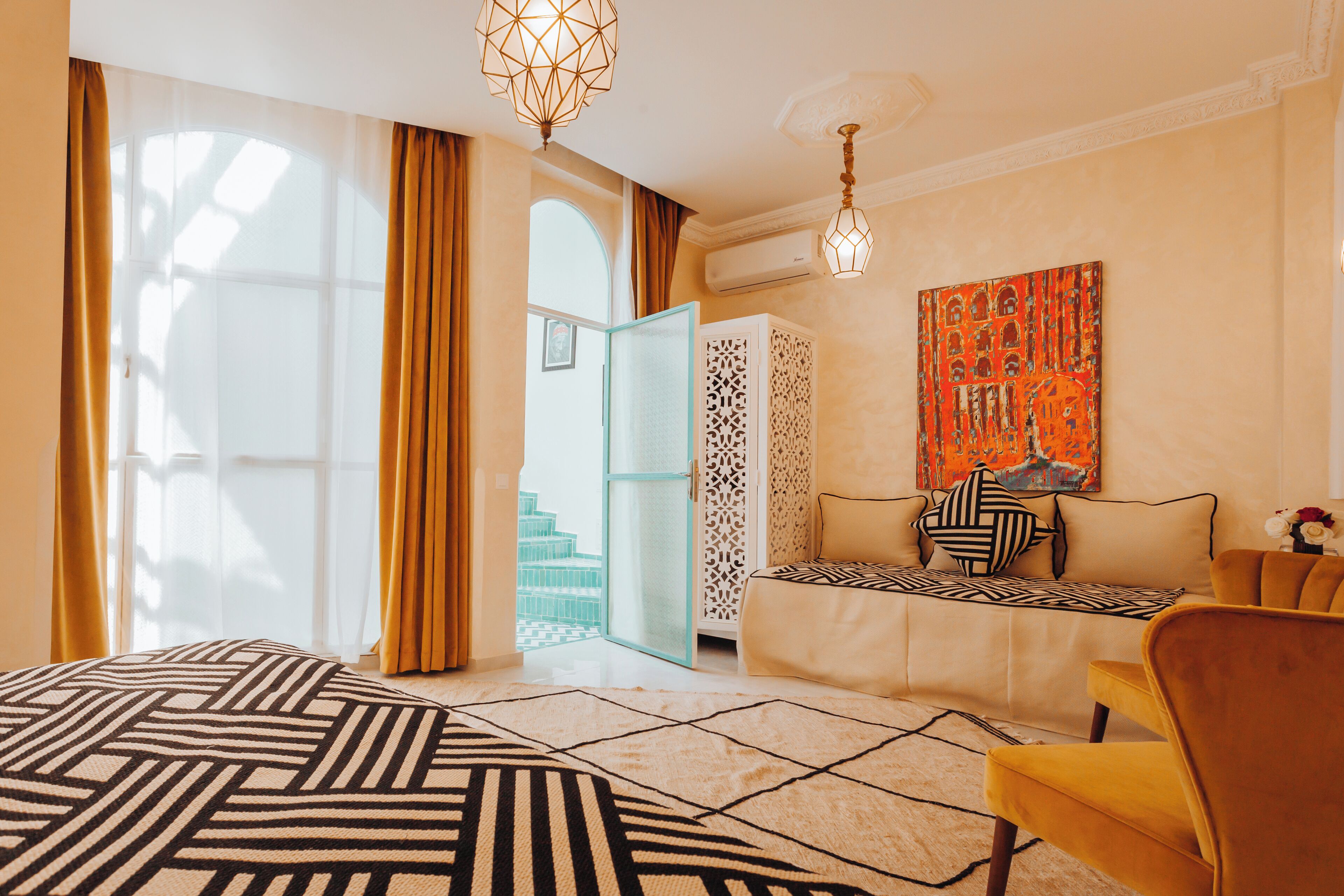 A hotel room with a bed, a sofa, a yellow armchair, a patterned rug, and a large window with curtains.