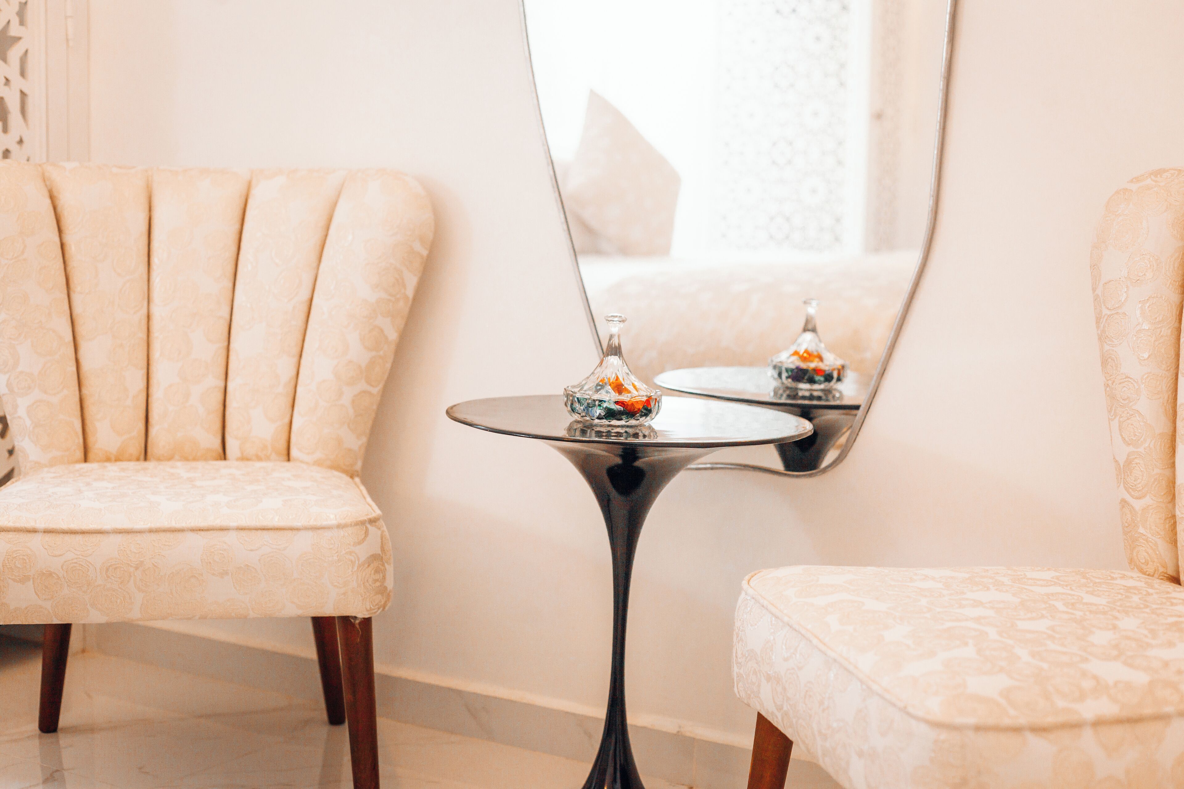 Une pièce comprenant deux fauteuils rembourrés, une petite table ronde avec un plateau en verre et un miroir qui reflète une partie de la pièce.