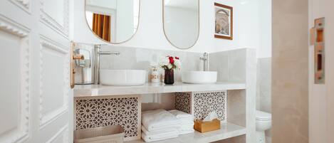 Un baño moderno con dos espejos ovalados grandes, un lavabo blanco y un inodoro. Hay azulejos decorativos en los estantes inferiores y un florero con flores en la encimera del lavabo.