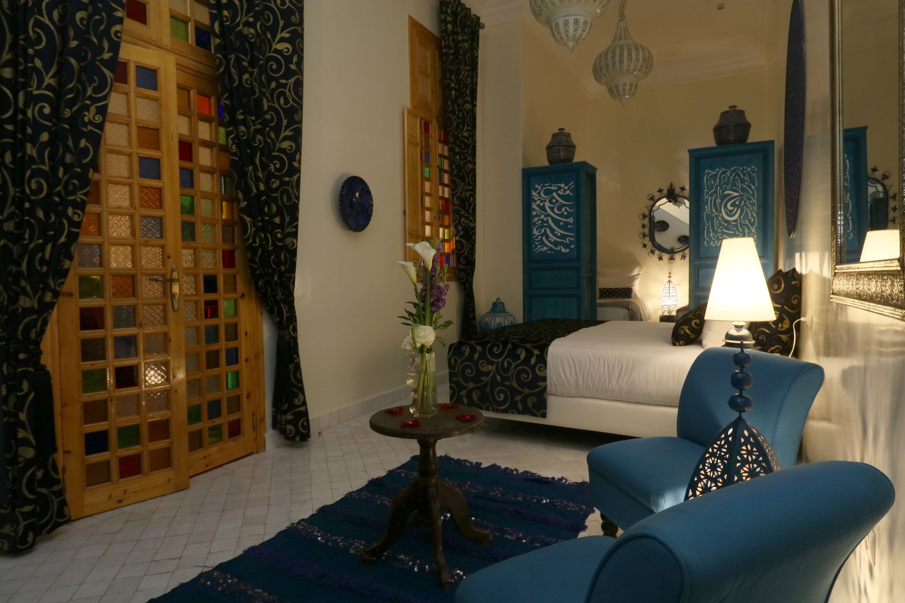 Une pièce comprenant un lit, un fauteuil bleu, une petite table avec un vase de fleurs et une porte en bois décorative.