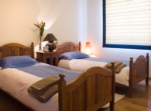 Two wooden beds with matching headboards, a bedside table with a lamp, a clock, and a vase with flowers. - Murteiras Apartment (Funchal)