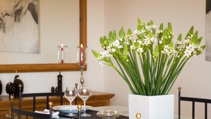 Et spisebord med en hvid vase med grønt løv og hvide blomster, to vinglas og et stearinlys på bordet.