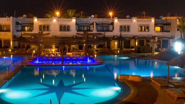 A swimming pool with a star-shaped light design, surrounded by lounge chairs and a building with arched windows and balconies.