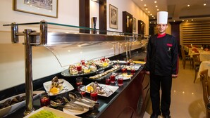 A buffet setup with various dishes and drinks, a waiter in uniform, and framed artwork on the wall.
