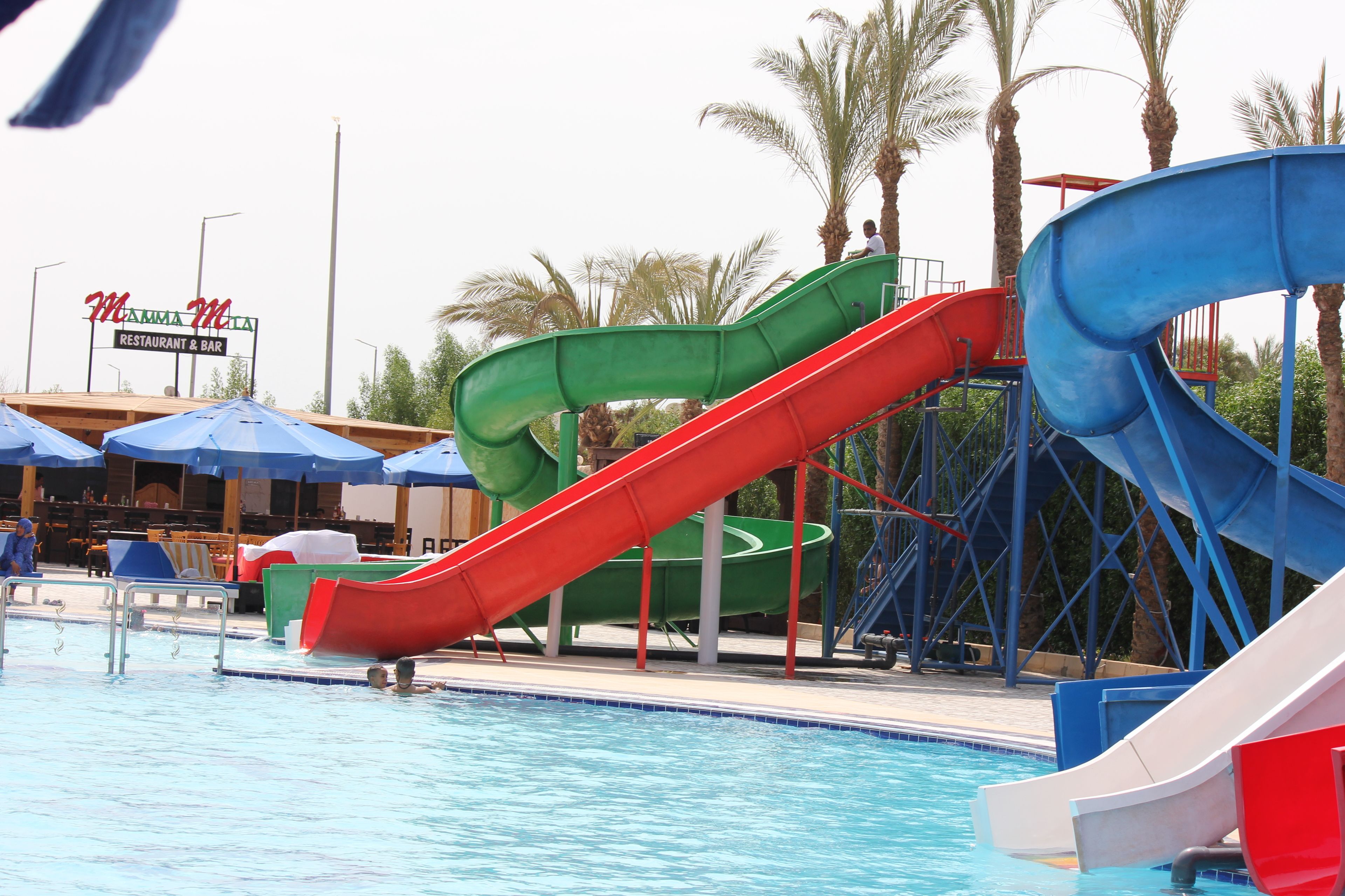 A pool area with multiple slides, a restaurant/bar sign, and palm trees in the background.