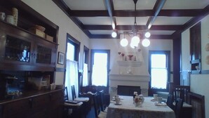 A dining room with a wooden table, chairs, and a fireplace. There are windows on the walls and a chandelier above the table.