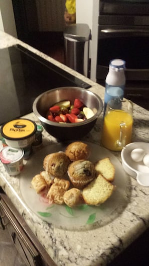 A breakfast setup with a bowl of fruit, muffins, biscuits, and a glass of orange juice on a marble countertop.