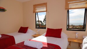 A hotel room with two beds, each with red bedding and white pillows, a small wooden nightstand, and a view of the sea through large windows. - Cabañas y Spa Costa Patagonia (Pichilemu)