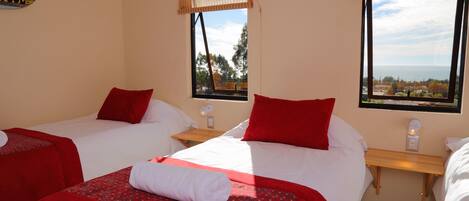 Habitación de hotel con dos camas, cada una con ropa de cama roja y almohadas blancas, una mesita de noche de madera y vistas al mar a través de amplias ventanas.