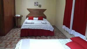 A bedroom with a bed, a wooden headboard, a small bedside table, and a window with red curtains. - Lagunillas del Poas (San Juan)