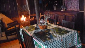 A rustic dining area with a stone fireplace, wooden chairs, and a table covered with a patterned tablecloth. - Lagunillas del Poas (San Juan)