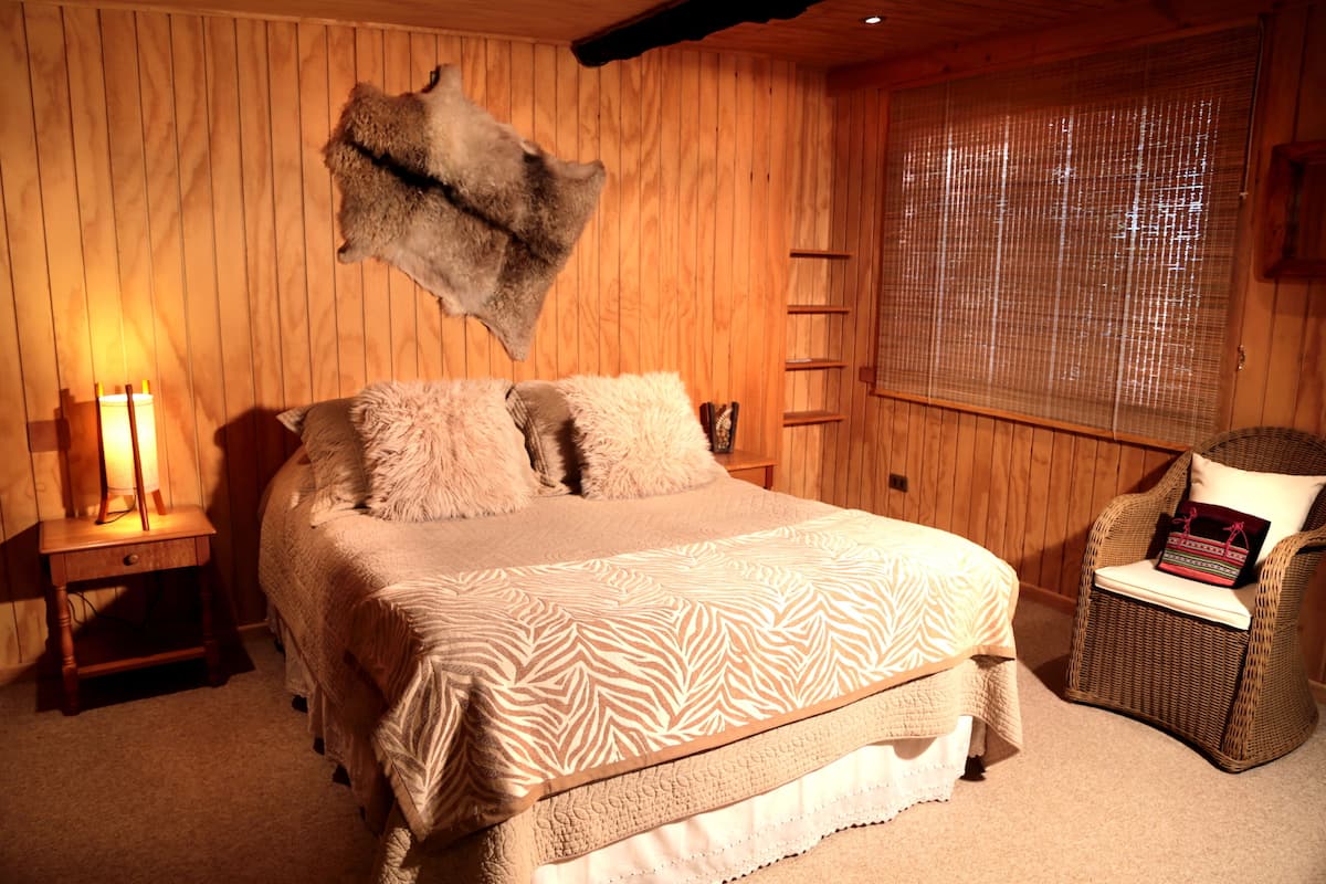 A wooden-paneled room with a bed, a chair, a wall-mounted fur piece, and a window with blinds.