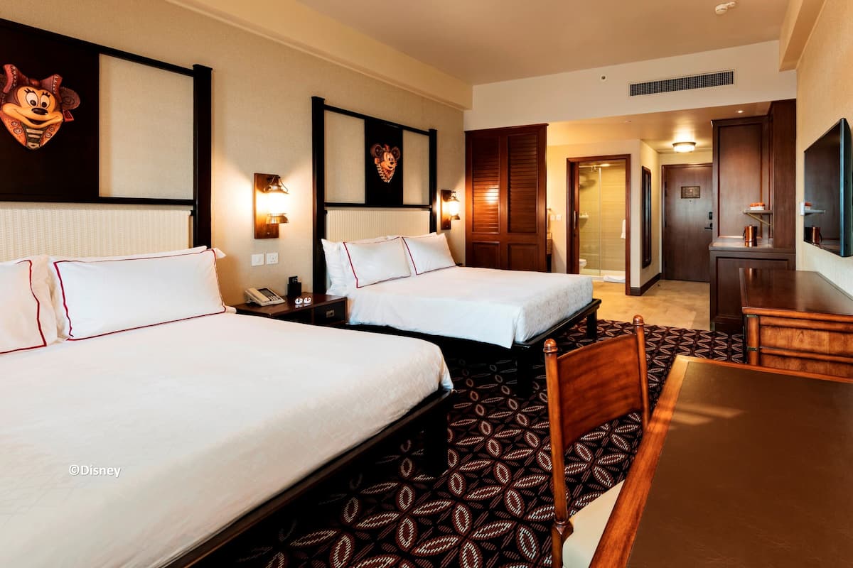 A hotel room with two beds, a desk, and a chair. There are wall-mounted lamps and a flat-screen TV.