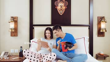 A hotel room with a bed, a bedside table with a phone and remote controls, and a wall-mounted Disney-themed artwork.