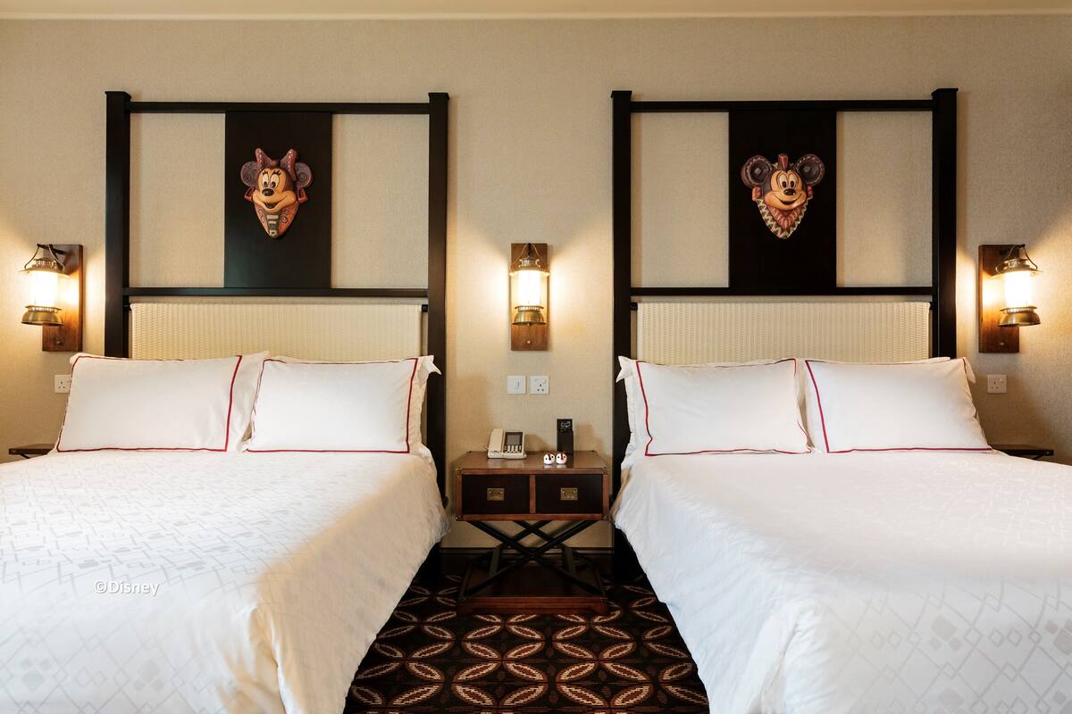 A hotel room with two beds, each with white bedding and red piping. There are wall-mounted light fixtures and a small table between the beds.