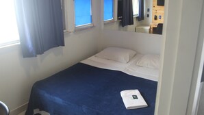 A single bed with a blue bedspread and white pillows in a room with a window and a mirror. - Maraca Hostel (Rio de Janeiro)