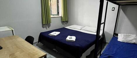 A dormitory room with two bunk beds, a single bed, a wooden table, and a window with curtains.