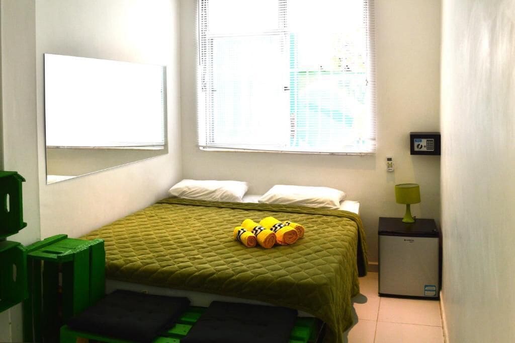A small, neatly arranged room with a bed, a desk, a lamp, and a window with blinds.