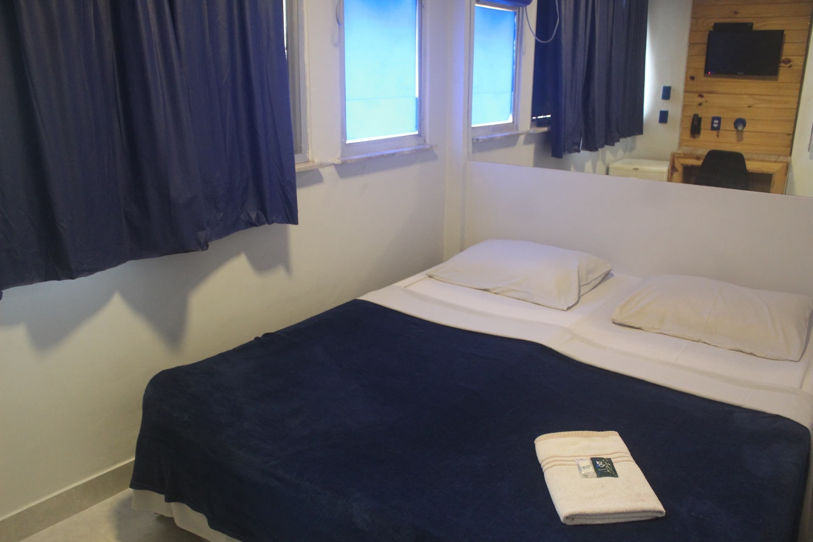 A single bed with a dark blue bedspread and two pillows, a folded towel on the bed, and a wooden desk with a chair in the corner.