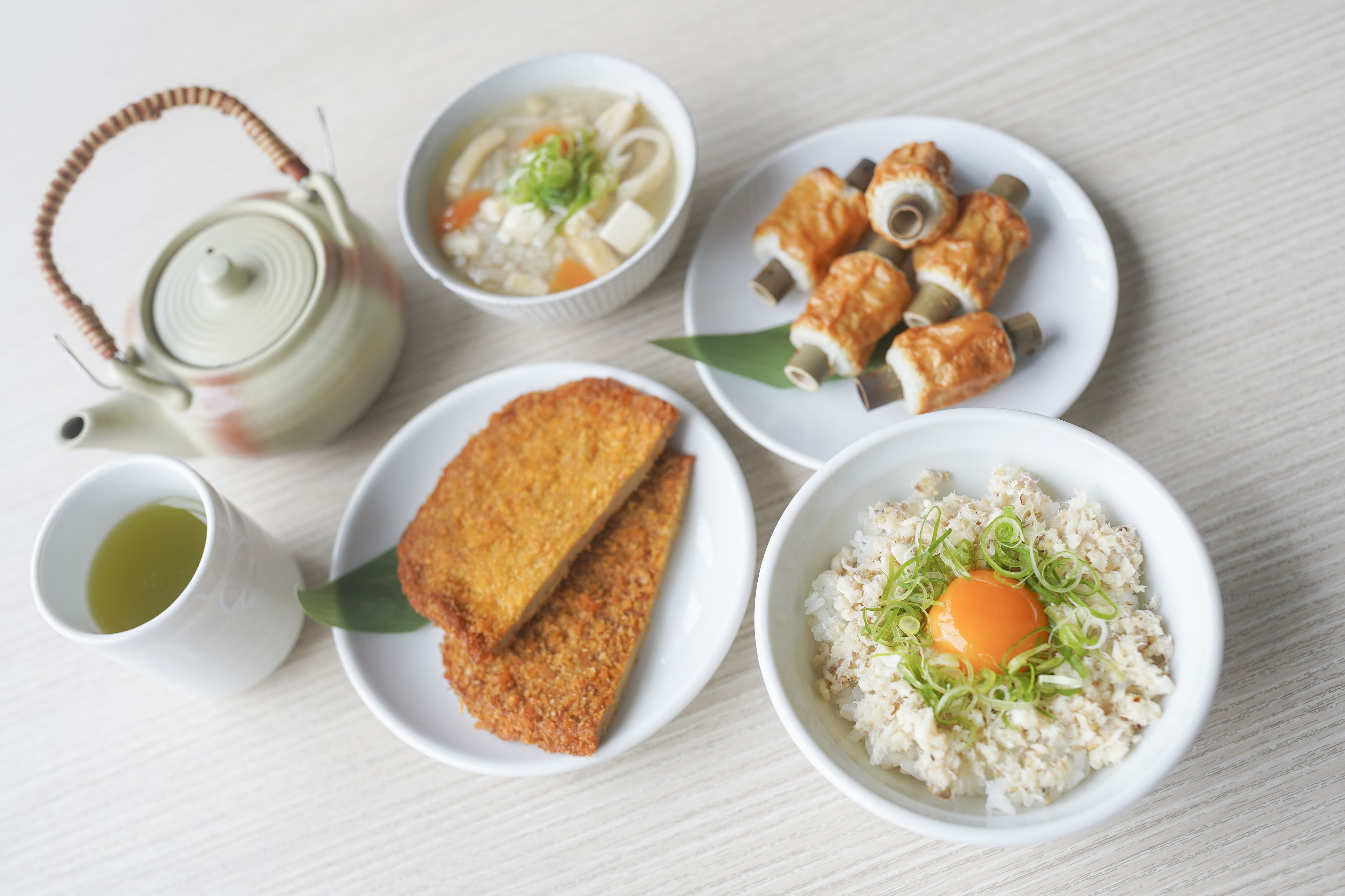 A table with various dishes including a teapot, a bowl of soup, a plate of tofu, a bowl of rice with an egg, and a cup of tea.