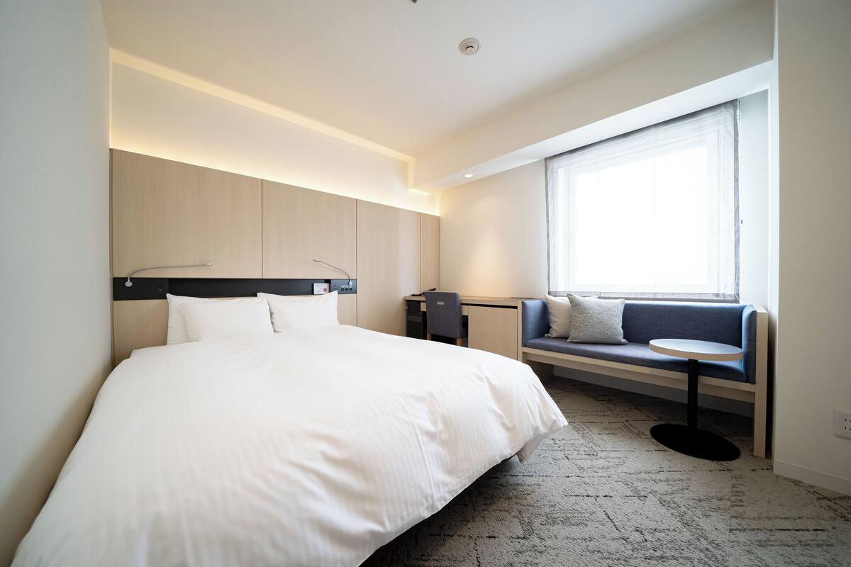 A modern hotel room with a large bed, a desk, a seating area with a sofa and a small table, and a large window providing natural light.