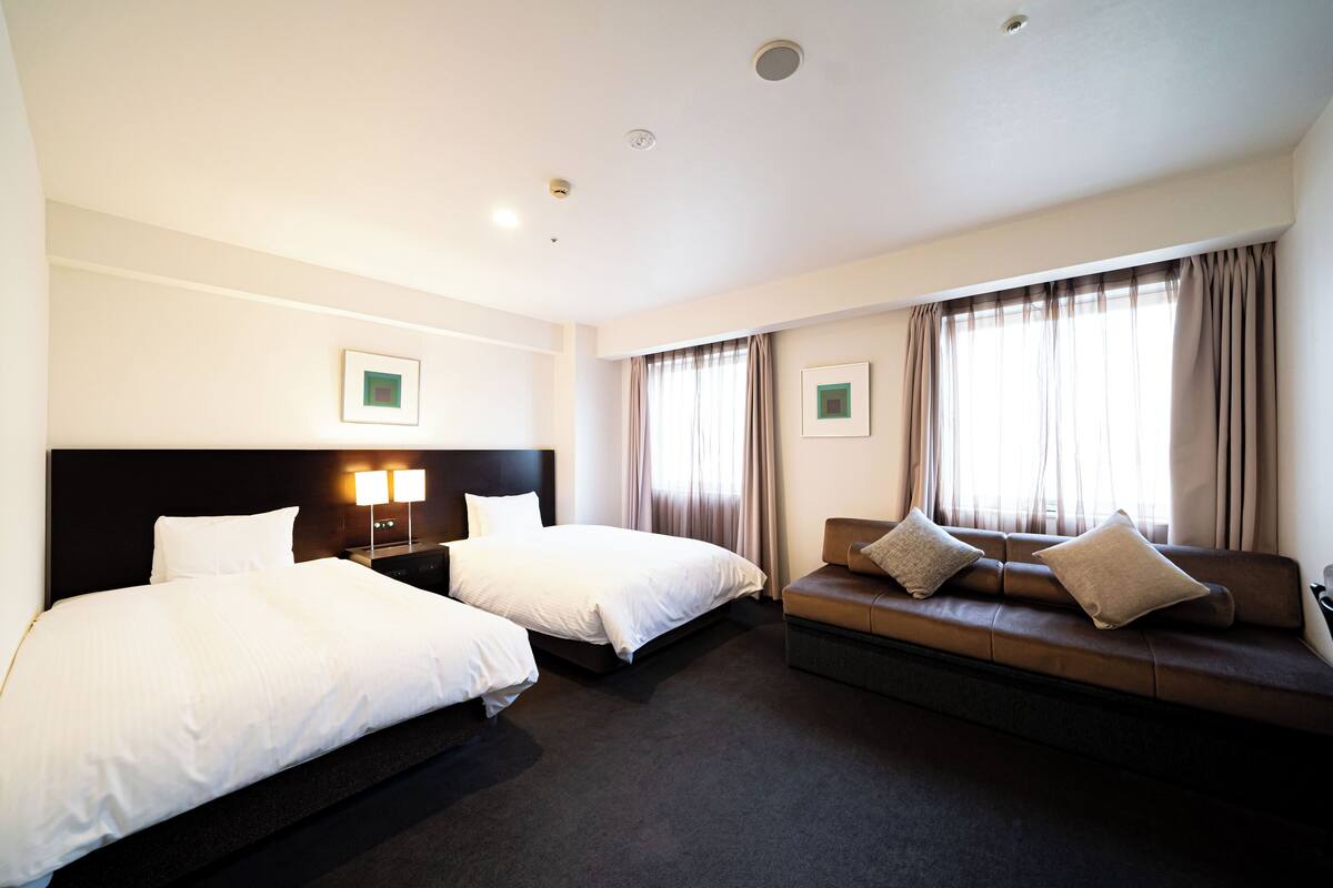 A hotel room with two single beds, a sofa, a nightstand with a lamp, and a window with curtains.
