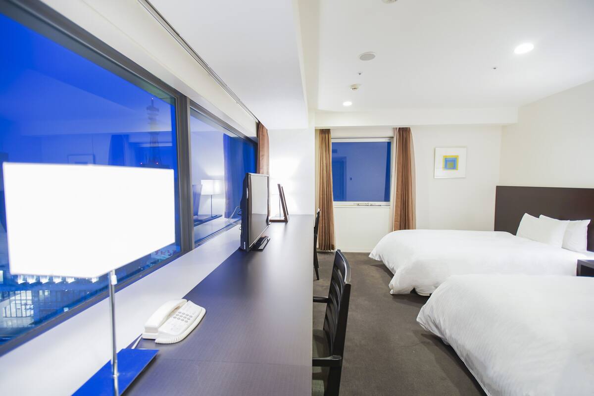 A hotel room with two beds, a desk with a telephone, a large window, and a view of the city.