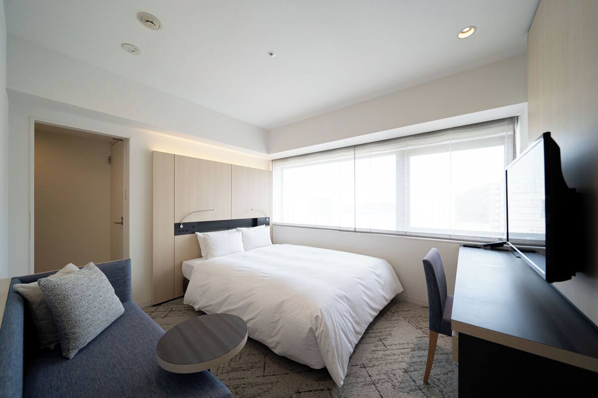 A modern hotel room with a bed, a desk, a chair, a sofa, and a TV.