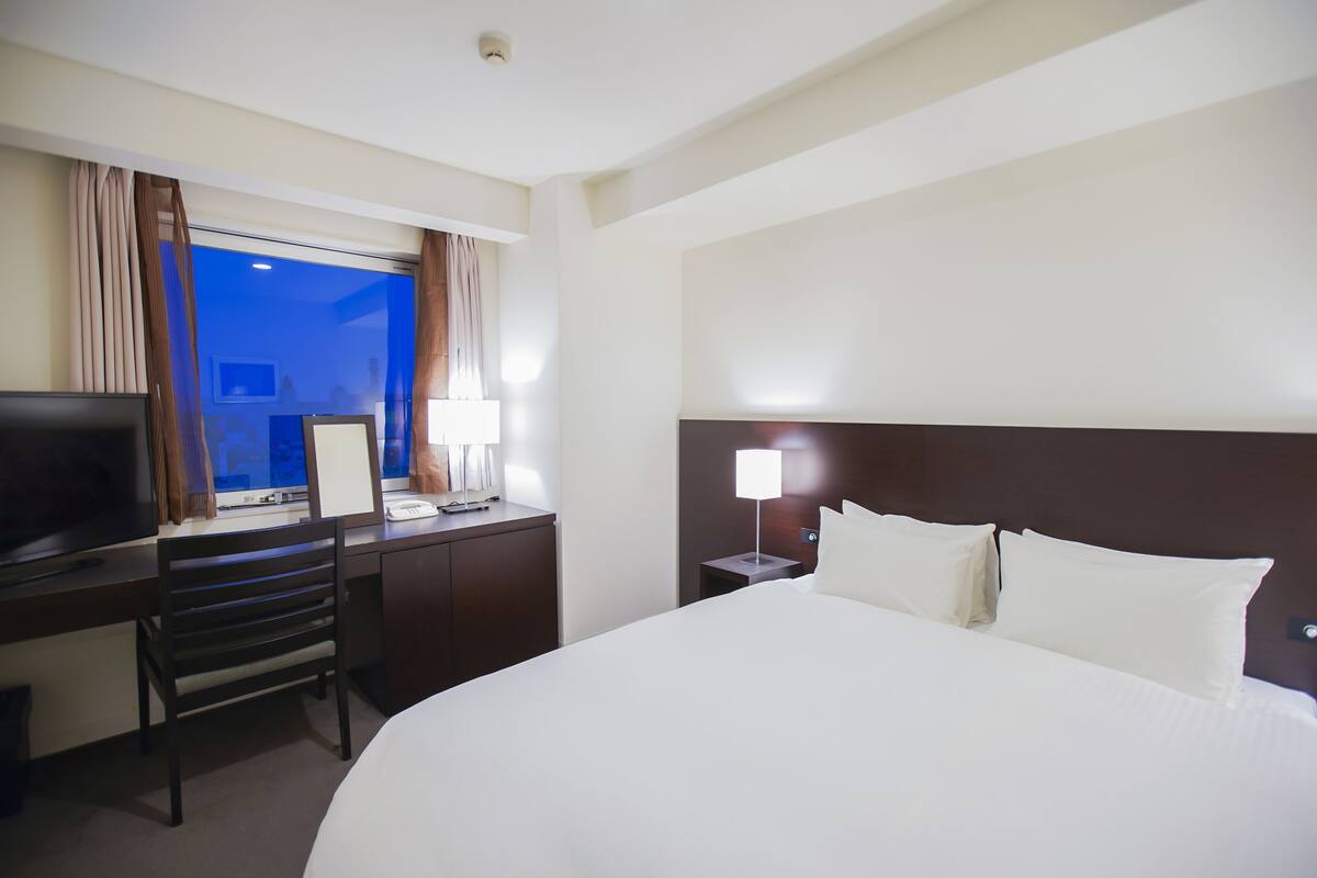 A hotel room with a large bed, a desk with a computer, a TV, and a window with curtains.