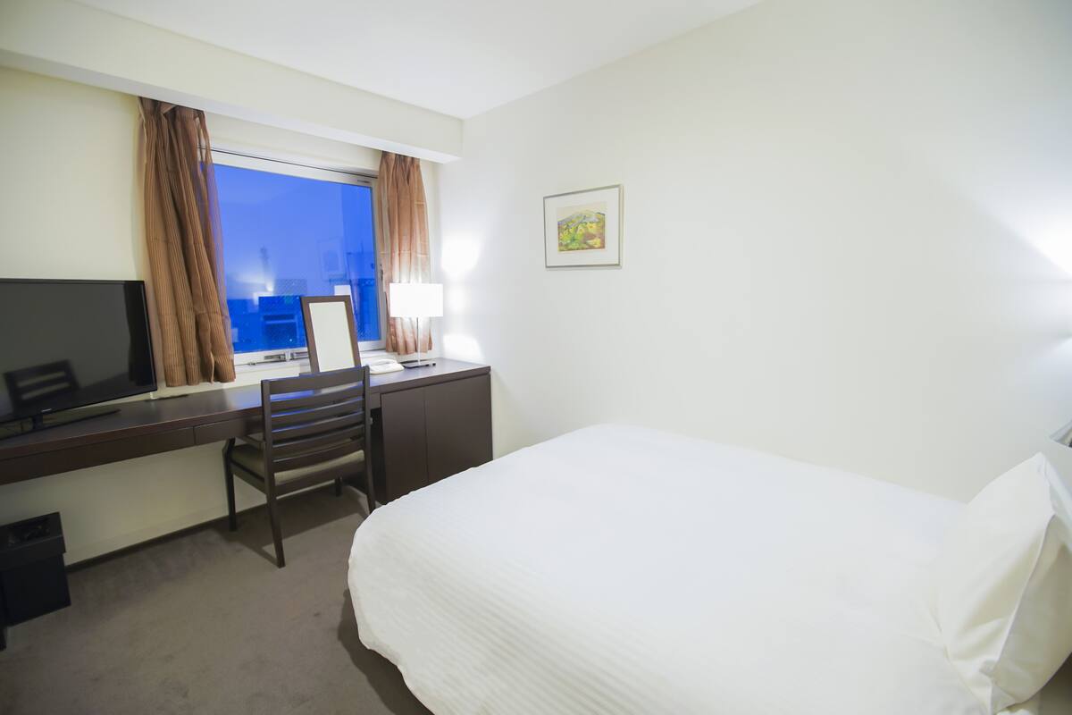 A hotel room with a bed, a desk with a chair, a television, and a window with curtains.