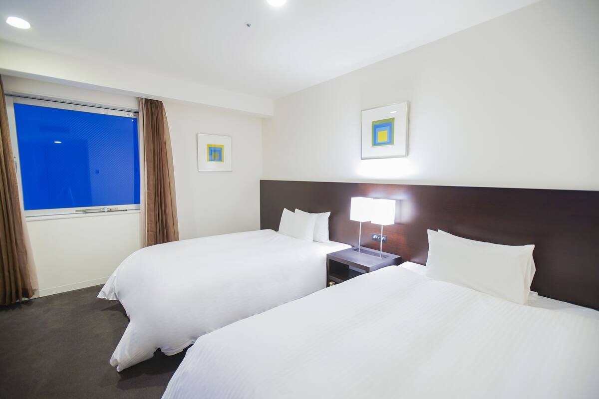 A hotel room with two beds, a window with curtains, and two framed pictures on the wall.