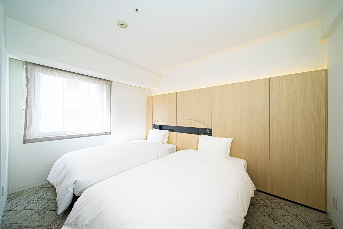 A hotel room with two beds, a large window, and wooden paneling on the wall.