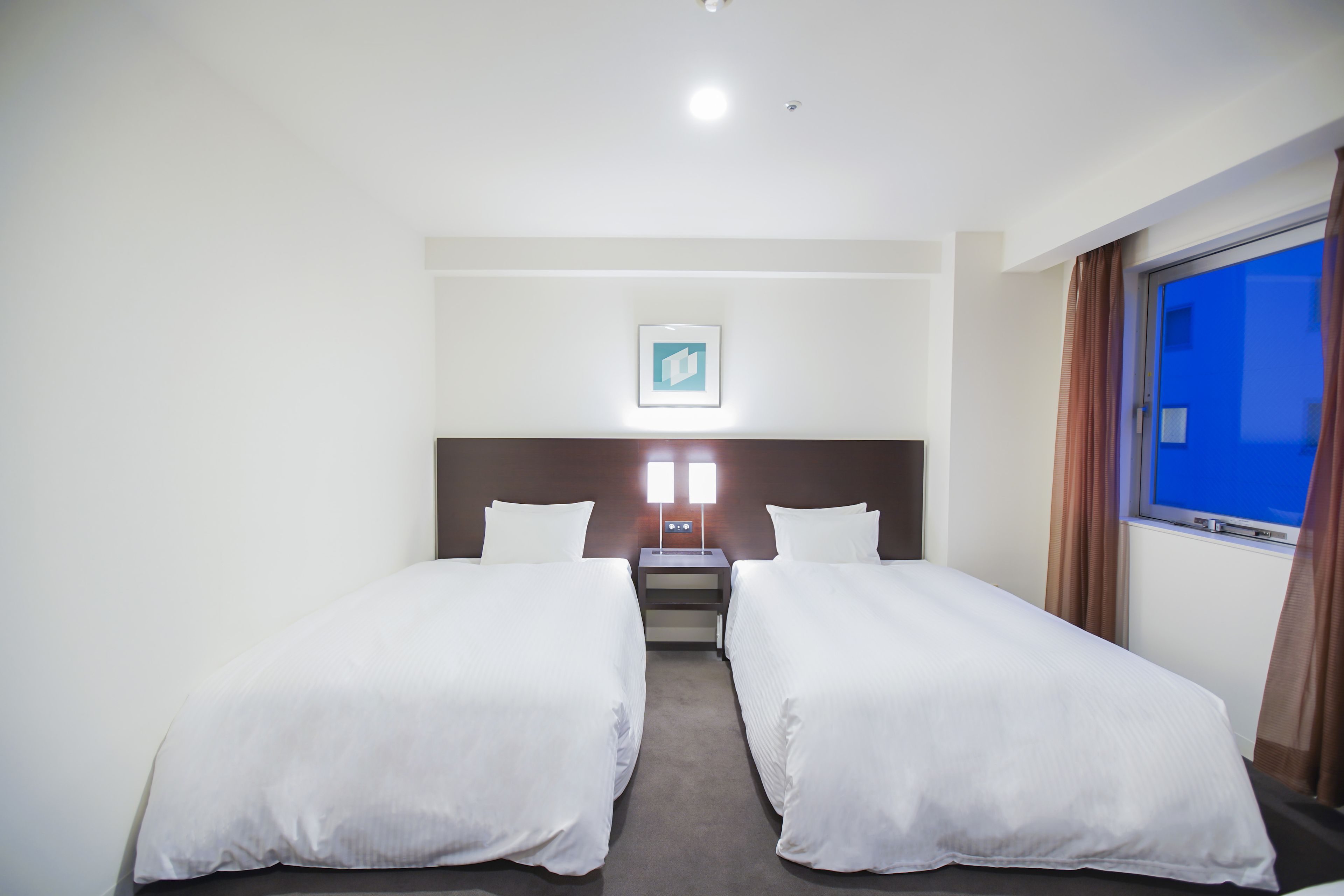A hotel room with two single beds, a nightstand, a framed picture on the wall, and a window with curtains.