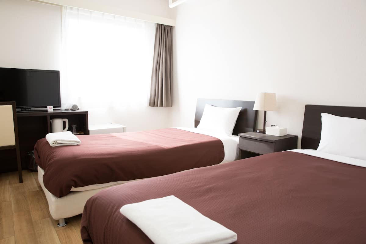 A hotel room with two beds, a TV, a desk, and a window with curtains.