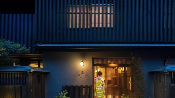 Een traditioneel Japans gebouw met een houten ingang, een bord met de tekst 'The Tenei Hotel', en een persoon in traditionele kleding die binnenstapt.