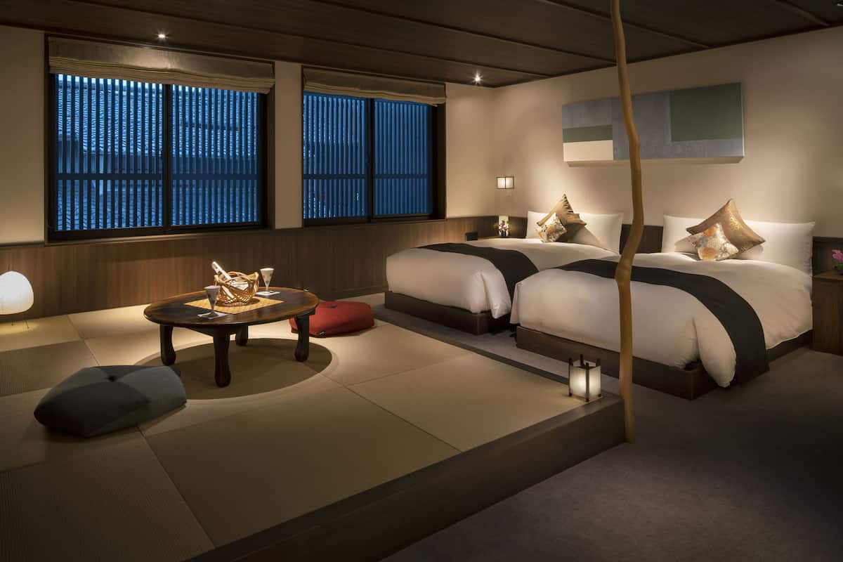 Ein Hotelzimmer mit Tatami-Boden, zwei Betten mit Kissen, einem kleinen Tisch mit Lampe und einem Fenster mit vertikalen Jalousien.