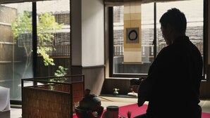 Een persoon zittend op een tatami mat in een kamer met een traditionele Japanse theeceremonieopstelling, inclusief een theebeker, een theecaddy en een bamboe mand.
